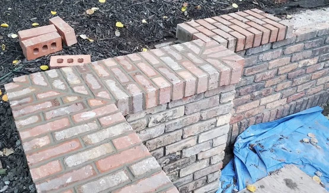 Brick wall construction in progress with a corner section showing newly laid red bricks, loose bricks nearby, and a blue tarp on the ground.