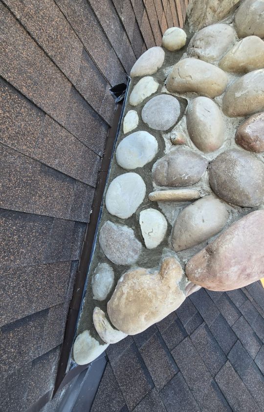 A section of a rooftop where brown shingles meet a stone chimney, with visible flashing between the materials.