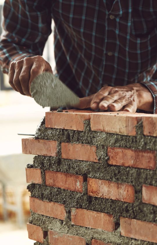 A person wearing a plaid shirt uses a trowel to spread mortar on a brick wall under construction.
