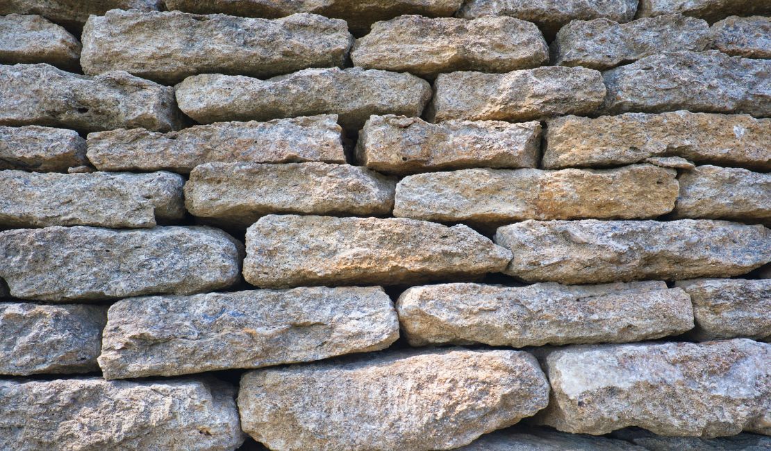 A close-up of a stone wall made of uneven, weathered rectangular rocks stacked in horizontal rows.