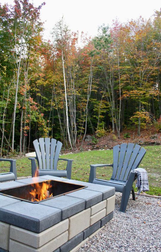 A square stone fire pit with a small fire burning, surrounded by four gray Adirondack chairs, set on gravel with autumn trees in the background.