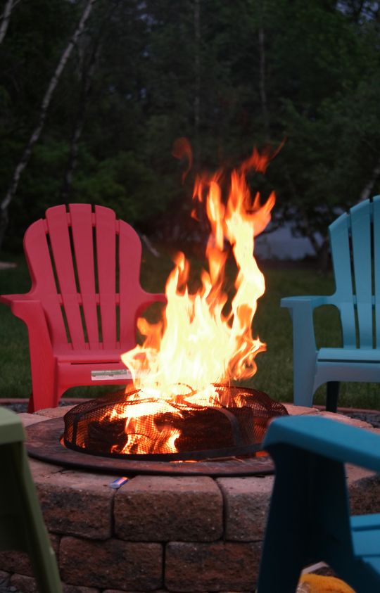 A bright campfire burns in a firepit surrounded by colorful Adirondack chairs on a grassy lawn.