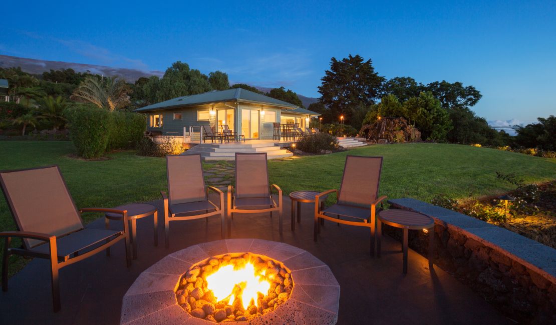 Five patio chairs arranged around a lit fire pit in a backyard, with a house and manicured lawn in the background at dusk.