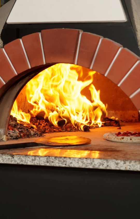 A wood-fired pizza oven with flames inside, baking a pizza next to burning wood and hot embers.