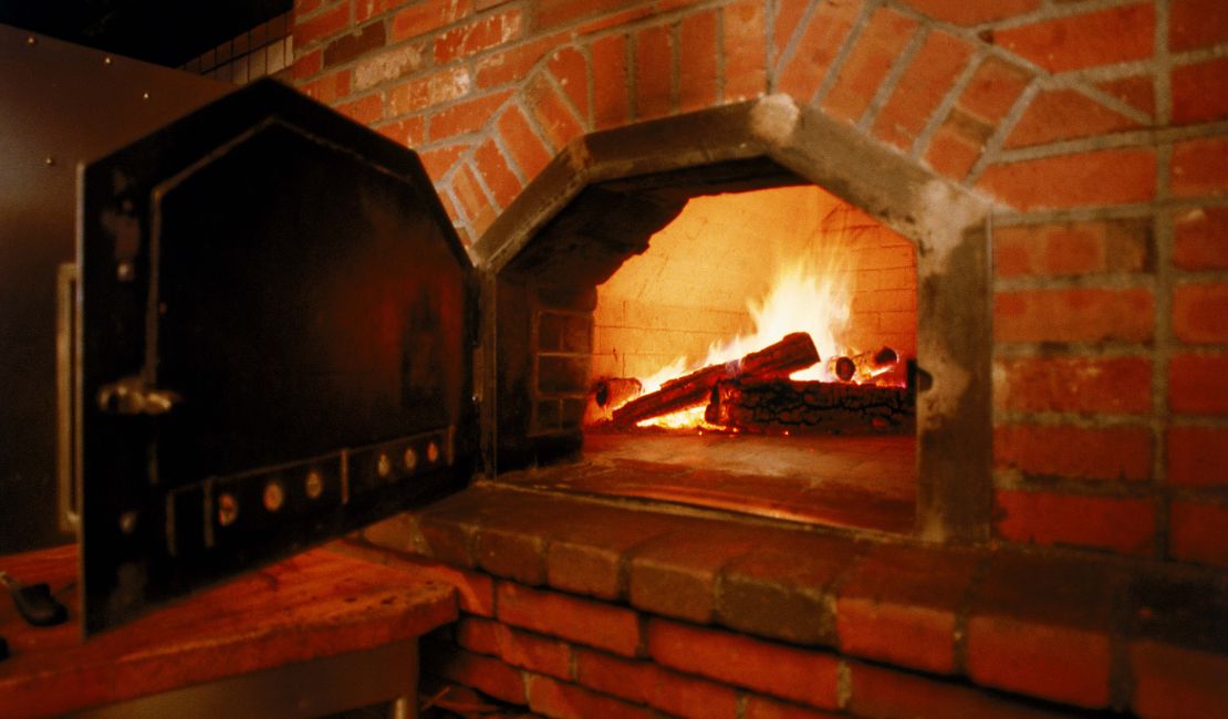 A wood-fired brick oven with an open black metal door and burning logs inside.