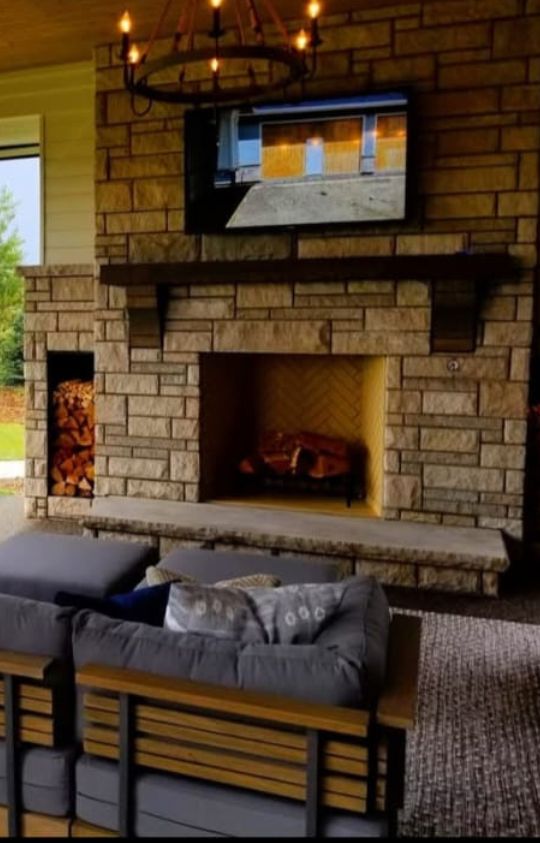 A cozy outdoor seating area with cushioned chairs facing a stone fireplace, topped by a mounted television and decorative chandelier.