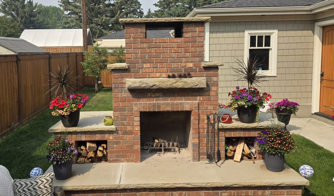 Outdoor brick fireplace with a wood storage area on each side, surrounded by colorful potted flowers on a patio beside a house.