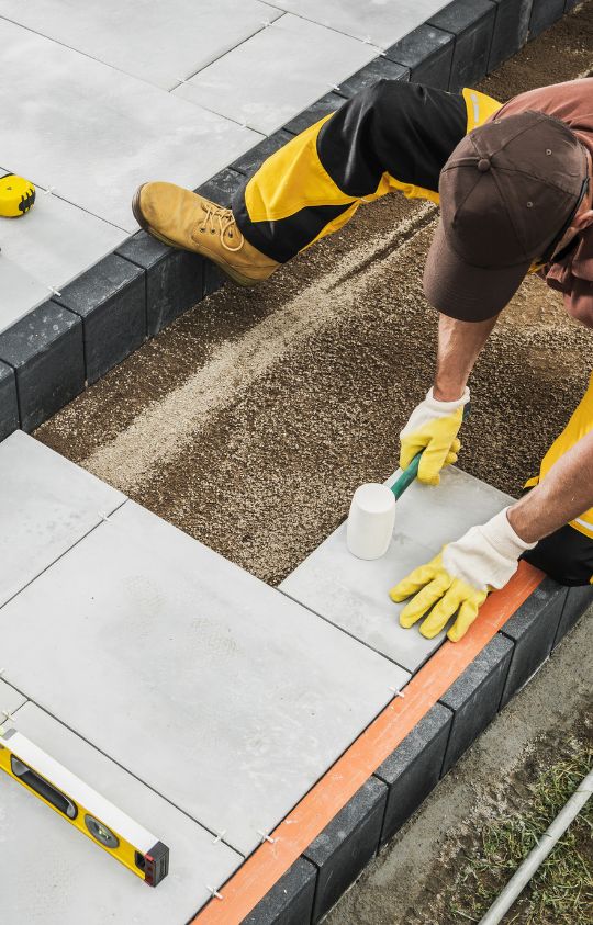 Worker in yellow gloves and uniform laying concrete paving slabs, using a rubber mallet and leveling tools to ensure proper alignment.