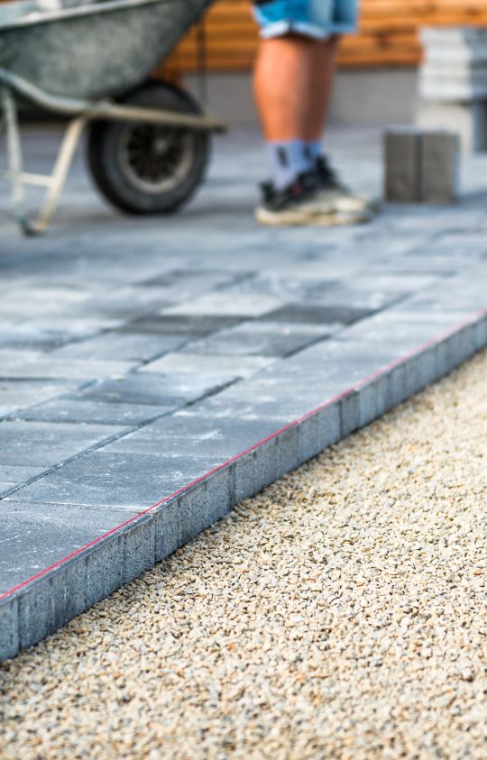 A worker stands next to a wheelbarrow while laying gray paving stones on a gravel base during outdoor construction.