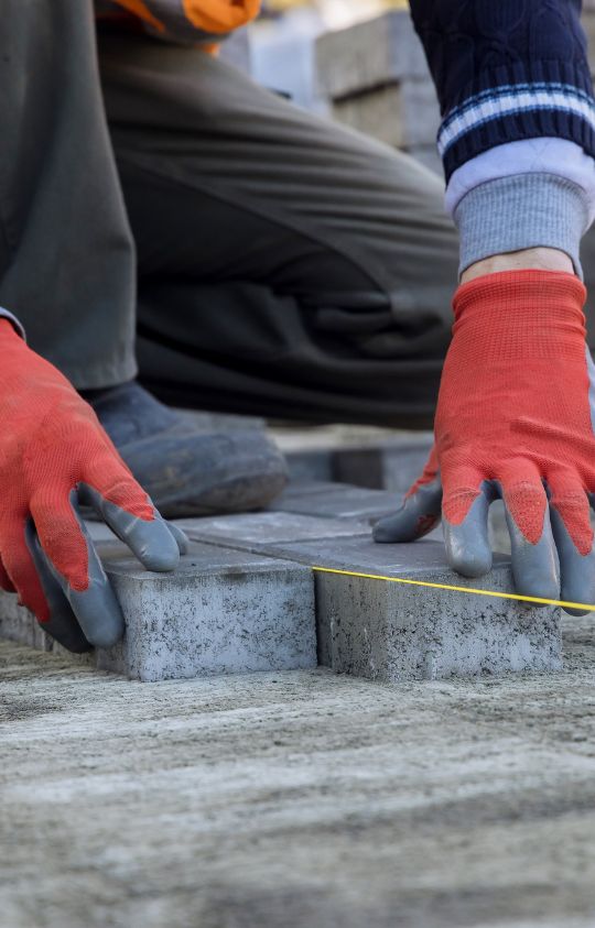 A worker wearing orange gloves carefully aligns concrete paving stones with a yellow measuring line.