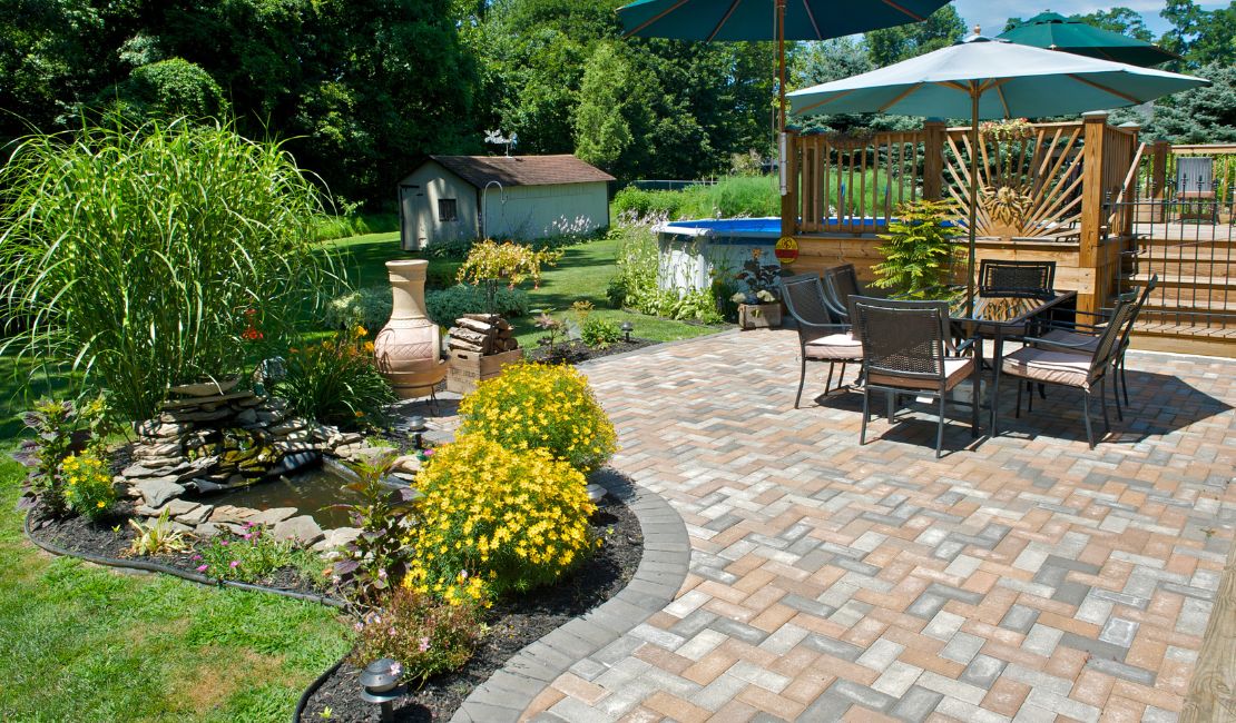 Backyard patio with outdoor dining set under large umbrellas, a small pond with yellow flowers, and a wooden deck near a pool.