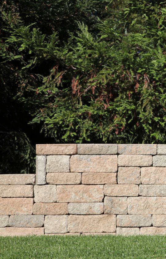 A low stone wall with blocks of light brown bricks stands in front of dense green foliage and a grassy lawn.