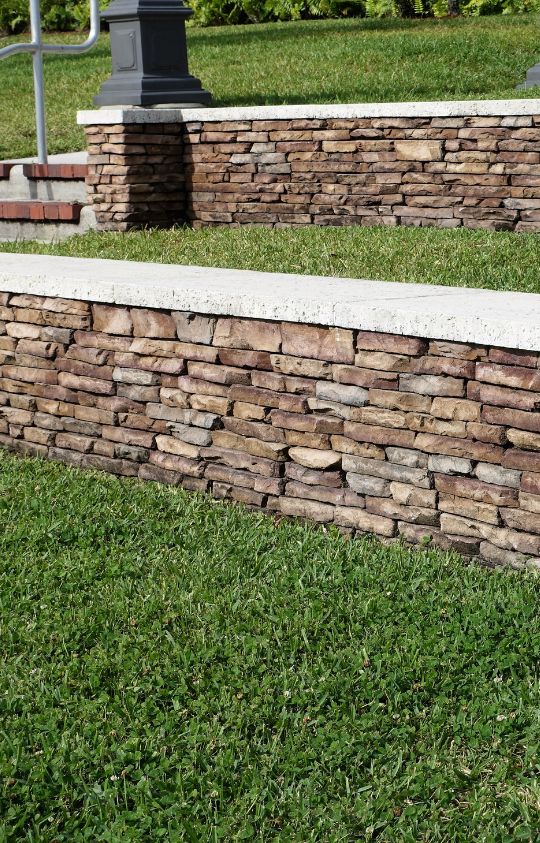Tiered stone retaining walls with concrete caps separate levels of green grass in an outdoor landscaped area.