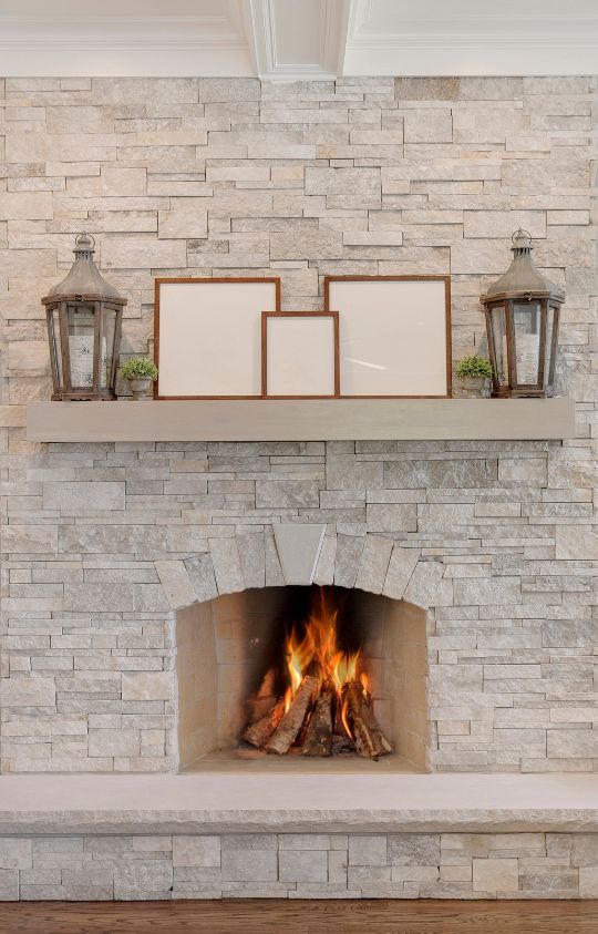 Modern stone fireplace with a lit fire, featuring a mantel decorated with lanterns, framed artwork, and small potted plants.