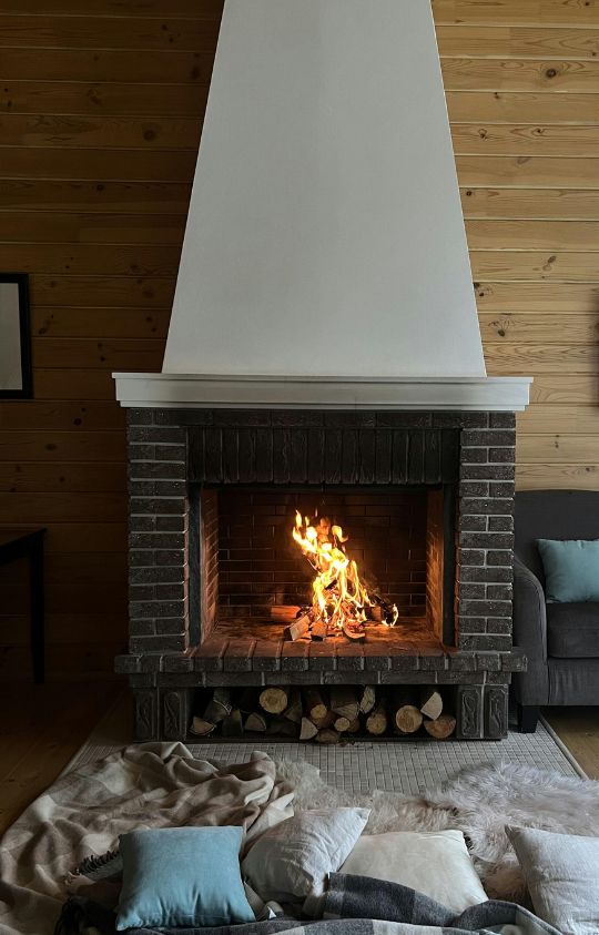 A cozy brick fireplace with a bright fire burning, surrounded by wooden walls, a gray sofa, and a blanket with pillows on the floor.
