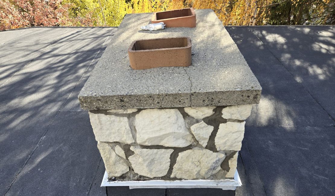 A stone chimney on a black flat roof with a concrete cap and two rectangular clay flue liners, showing signs of cracked mortar.