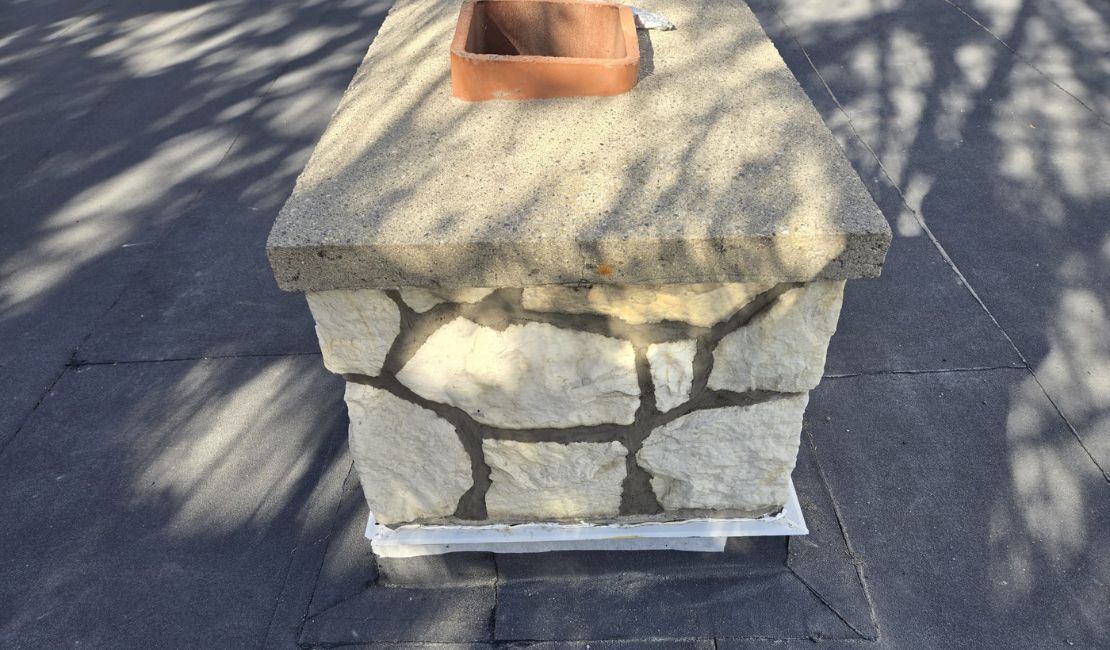 A rectangular chimney with a concrete top and stone sides sits on a flat roof, casting shadows in the sunlight.