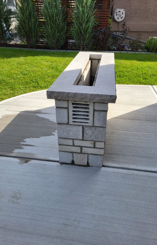 custom outdoor gallery potrait 1 Rectangular stone fire pit with vent, sitting on a concrete patio surrounded by a green lawn and shrubbery.