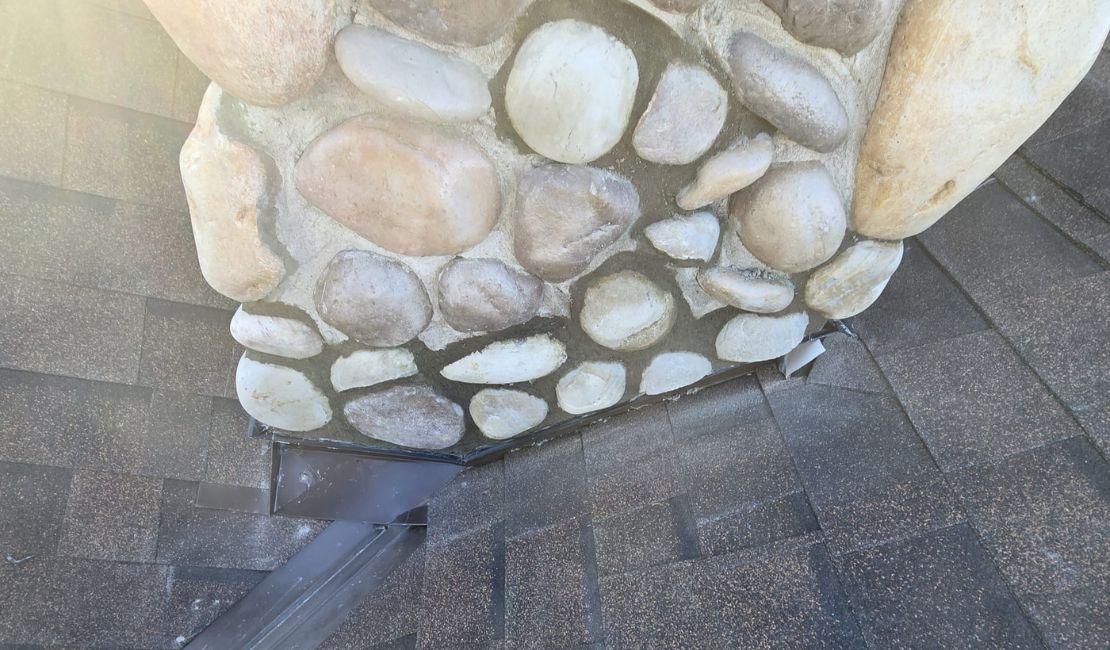 Close-up of a stone chimney with visible metal flashing installed at the base, surrounded by brown roof shingles.