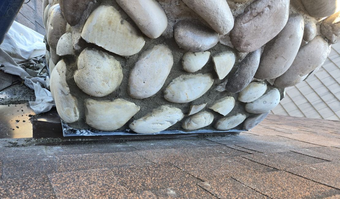A close-up view of a stone chimney base on a shingled roof with metal flashing and some construction materials nearby.
