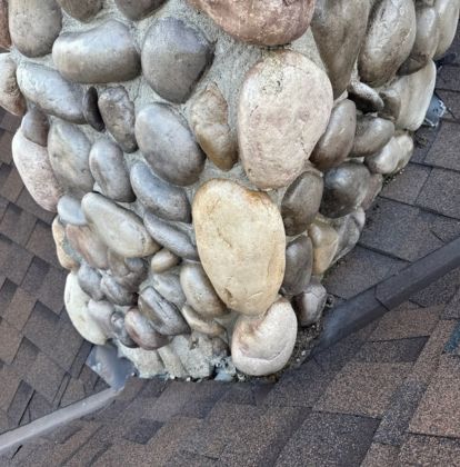 Stone chimney with rounded rocks extending from an asphalt shingle roof, with visible flashing around the chimney base.