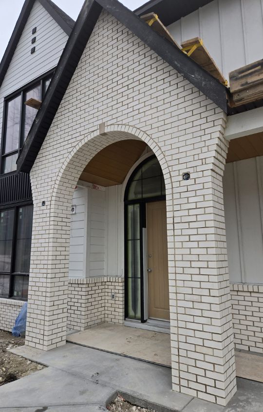A modern house entrance with a tall arched brick doorway, light wood door, and vertical siding on the exterior walls.