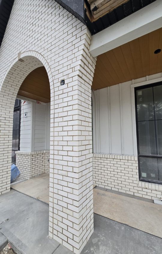 Brick archway and column on a porch with a combination of white brick and vertical white siding, unfinished entryway.