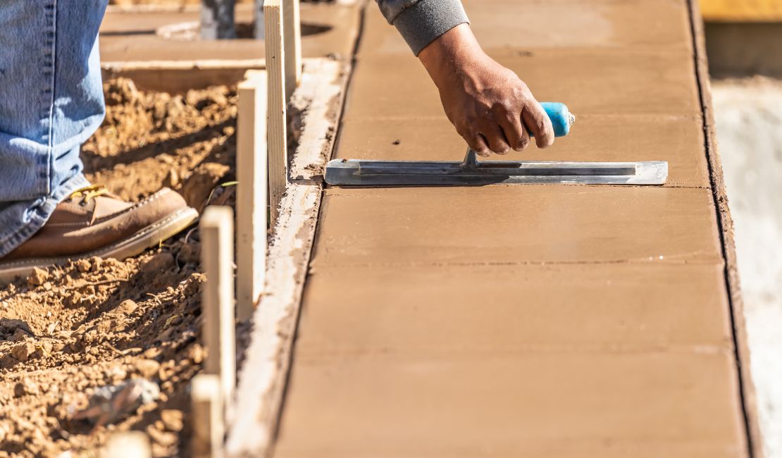 A worker smooths wet concrete on a newly constructed sidewalk using a hand trowel, with wooden forms on the side.
