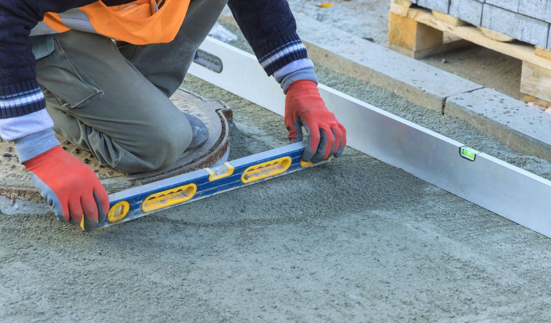 A construction worker uses a spirit level to check the evenness of freshly poured concrete while wearing red gloves.