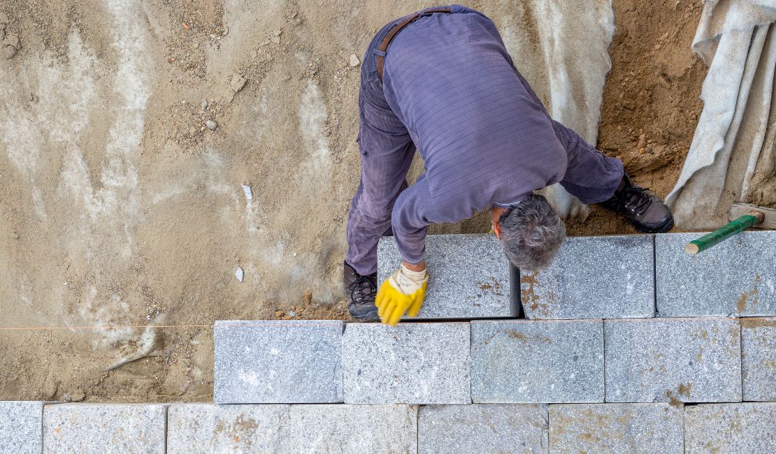 A worker wearing gloves lays gray stone bricks in a row on a sandy surface, viewed from above.
