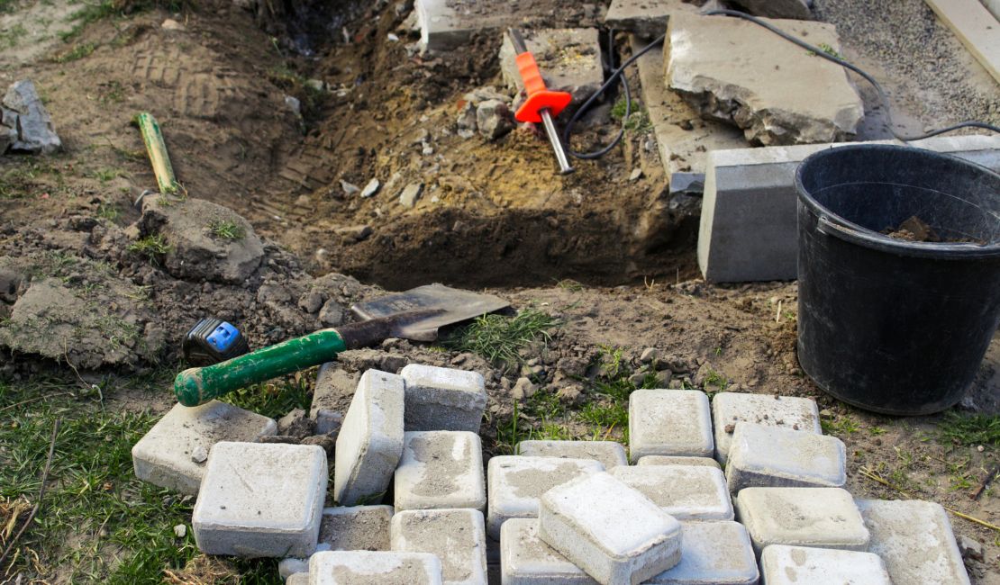Paving stones, tools, and a black bucket scattered on the ground at a partially dug construction or landscaping site.