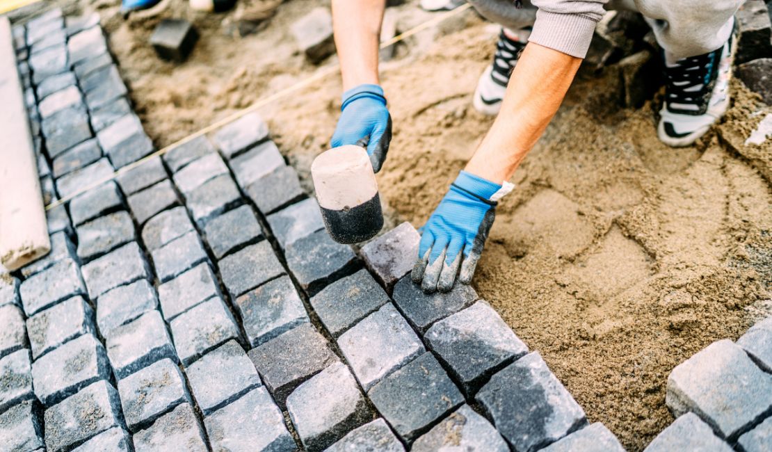 A person wearing blue gloves uses a rubber mallet to lay stone pavers on sand for a walkway or patio.