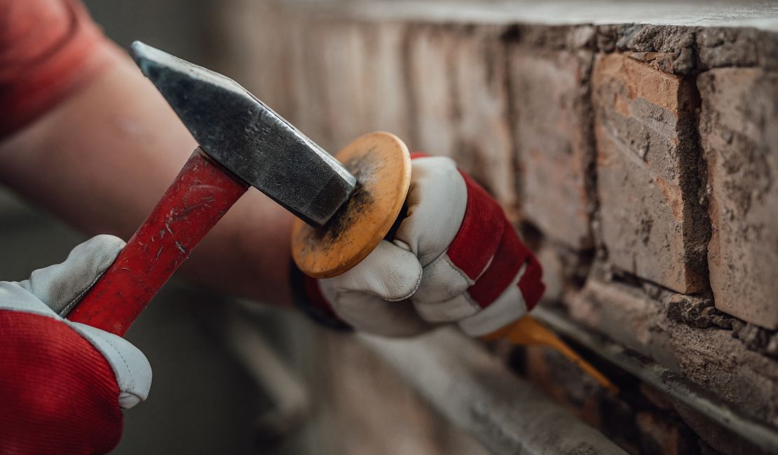A person wearing work gloves uses a hammer and chisel to remove old mortar between bricks on a wall.