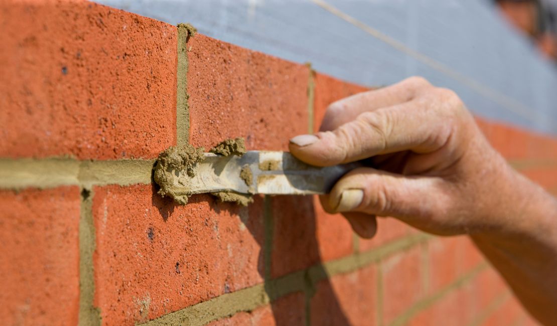 A hand uses a trowel to apply mortar between red bricks on a brick wall.
