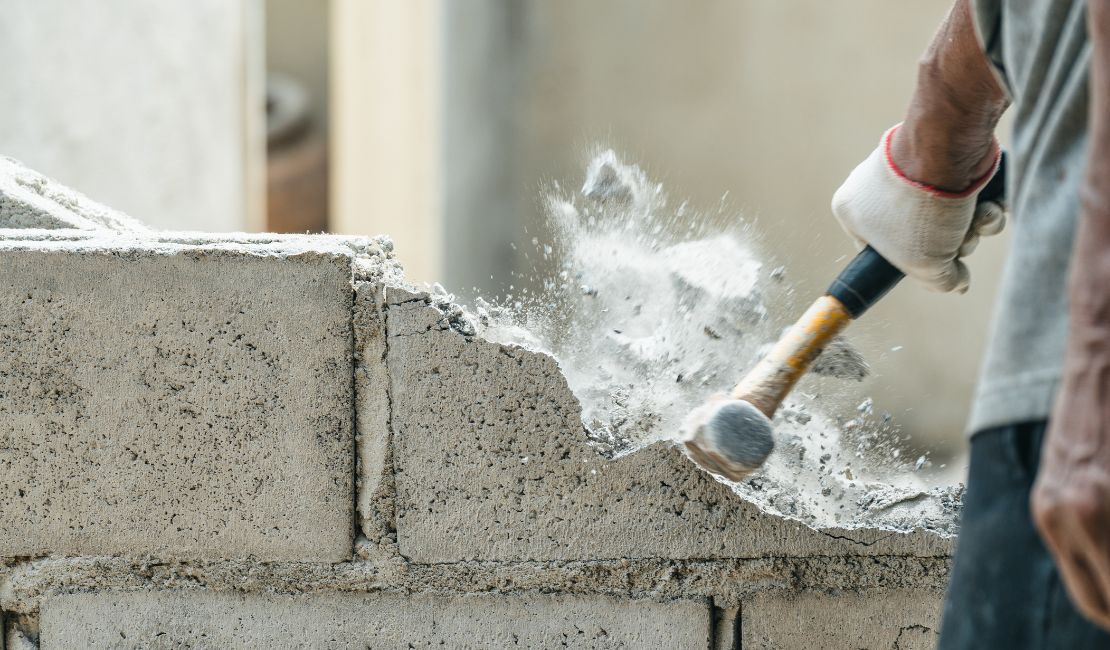 A person wearing gloves breaks a concrete block wall with a sledgehammer, causing dust and debris to fly.