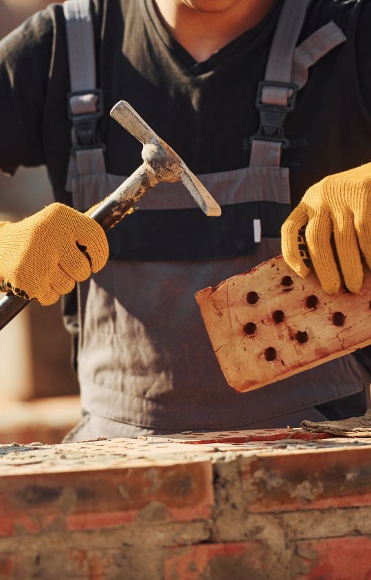 A construction worker wearing overalls and yellow gloves holds a brick in one hand and a masonry hammer in the other.