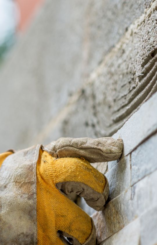 A gloved hand carefully placing or adjusting a stone tile on a wall during construction or masonry work.