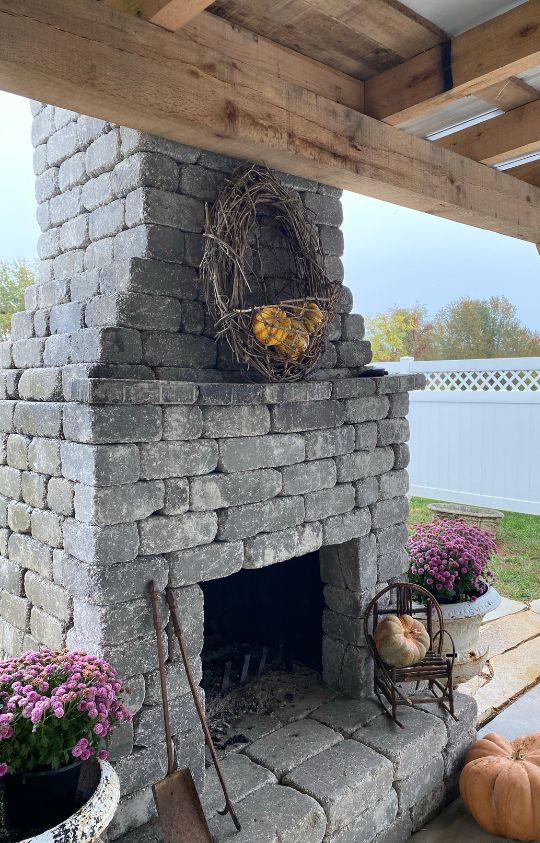 Outdoor stone fireplace decorated with a grapevine wreath holding yellow gourds, surrounded by pumpkins and potted pink flowers.