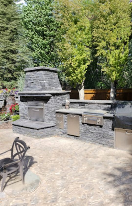 Outdoor stone kitchen with a built-in fireplace, grill, and metal cabinets, set on a patio with trees and a wooden fence in the background.
