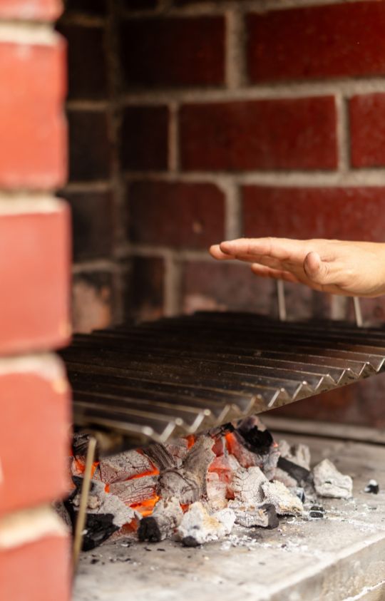 A hand hovers over a metal grill above hot coals in a brick barbecue.