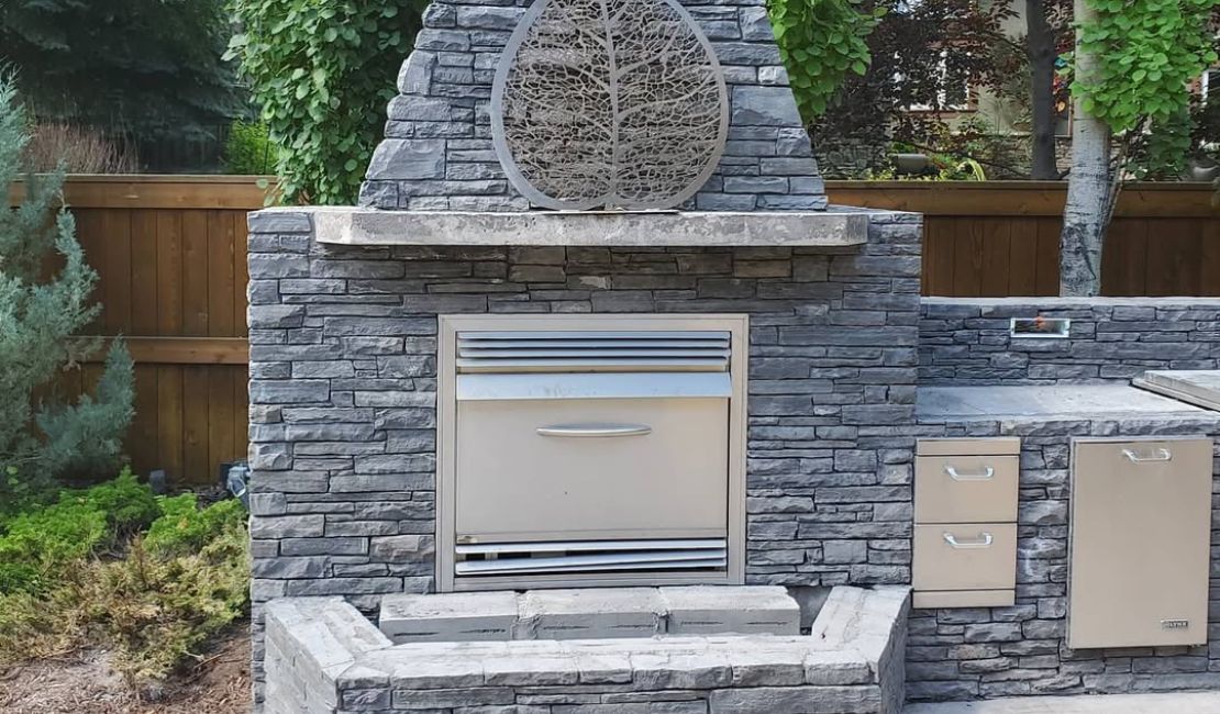 Outdoor kitchen with a built-in grill and stone surround, featuring a decorative metal leaf sculpture above the cooking area.