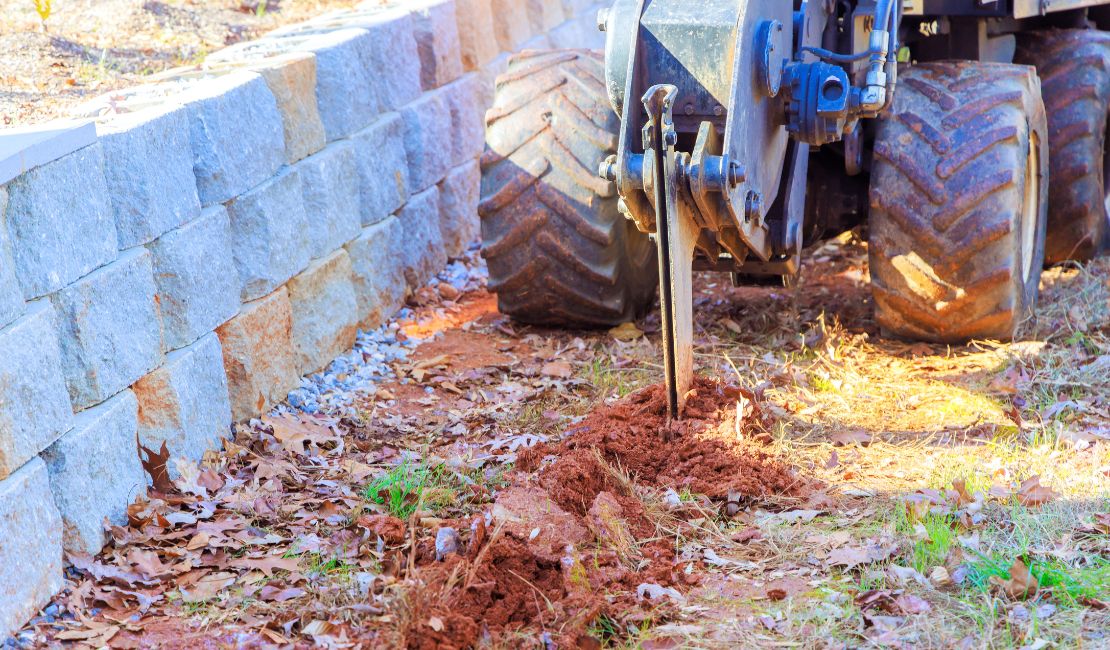 A small trencher machine digging a narrow trench beside a stone retaining wall on a patch of dry, leaf-covered ground.