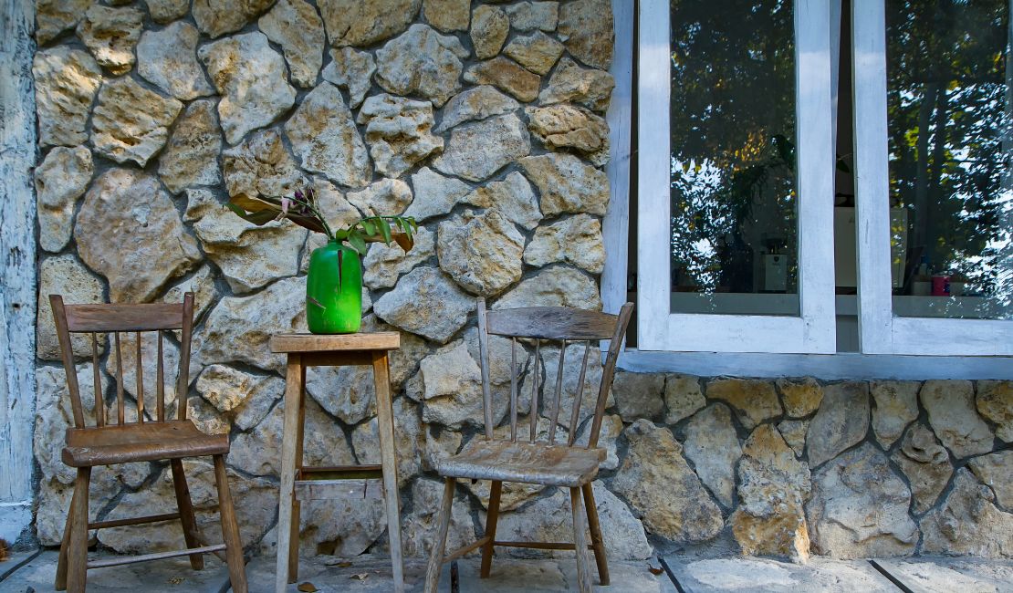 Two rustic wooden chairs and a small table with a green vase sit outside against a stone wall near a window.